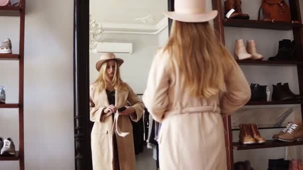 Vonzó szőke lány előtt álló a tükör bézs kabát és trendi kalap, hogy egy fotót nézni vele okostelefon. Élvezi a stílusát. Felvételeket a hátoldalon — Stock videók