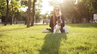 Çimenlerin üzerinde güneş gözlüğü takan, küçük köpeğini okşayan ve gülümseyen neşeli kızıl başlı bayanın ön görüntüleri. Yeşil çim, arka planda mercek fişekleri