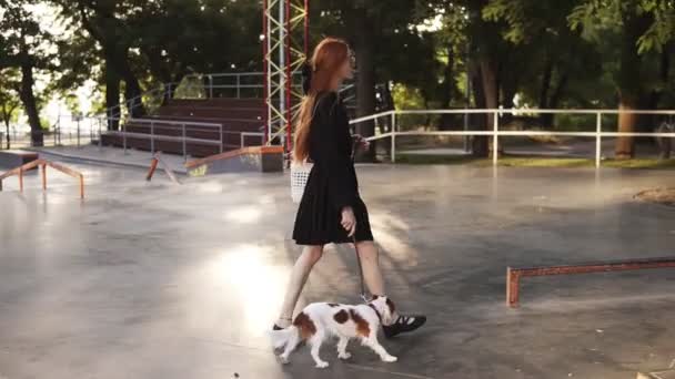Młoda, stylowa czerwona dziewczyna Chodzona przez pusty skatepark z kochającym psem. Powolny ruch Ładna dziewczyna w czarnej sukni chodzeniu czystorasowego psa w parku miejskim, rozglądając się. Widok z boku — Wideo stockowe