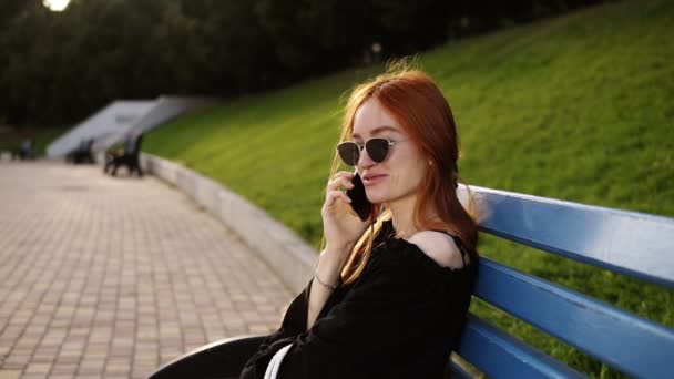 Портрет улыбающейся белой женщины с рыжими волосами, разговаривающей по мобильному телефону во время отдыха на скамейке в летнем парке. Веселая красивая женщина звонит по мобильному телефону, расслабляясь на открытом воздухе. Медленное движение — стоковое видео