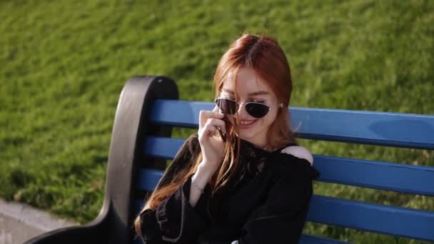 Retrato de mulher caucasiana sorridente com cabelo ruivo falando no celular enquanto descansa no banco no parque de verão. Mulher bonita alegre chamando no telefone celular enquanto relaxa ao ar livre. Movimento lento — Vídeo de Stock