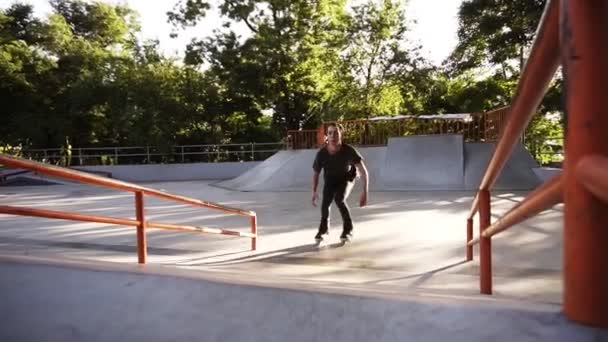 Встроенный роликовый фигурист делает трюки в бетонном скейтпарке на открытом воздухе с красивым зеленым фоном парка, замедленная съемка. Молодой человек проводит досуг в скейт-парке. Низкий угол съемки — стоковое видео