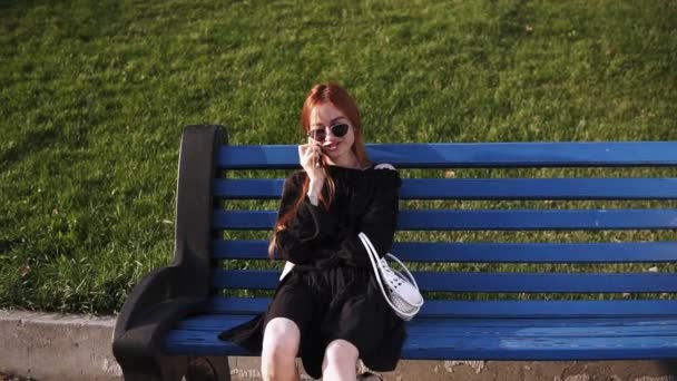 Портрет улыбающейся белой женщины с рыжими волосами в солнцезащитных очках и черным платьем, разговаривающей по мобильному телефону во время отдыха на скамейке в летнем парке. Веселая красивая женщина звонит по мобильному телефону в то время как — стоковое видео