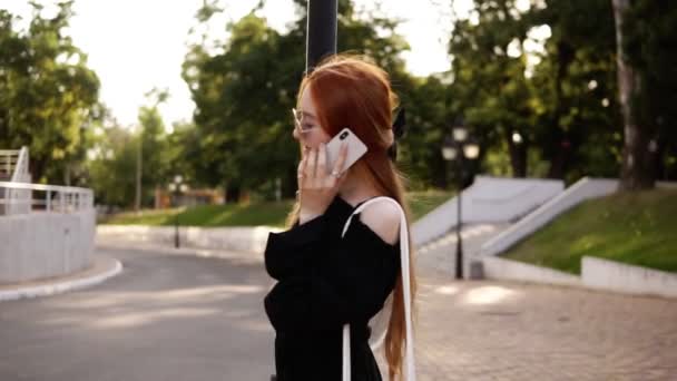 Jeune femme élégante en robe noire et lunettes de soleil parlant sur le téléphone portable tout en tournant autour du lampadaire dans le vert, parc de la ville — Video