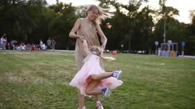 Küçük kızıyla bej elbiseli beyaz anneyi sevmek ve parkta oyun oynamak. Anne kızını döndürüyor, onlar mutlu ve publiv, yeşil parkta gülüyor