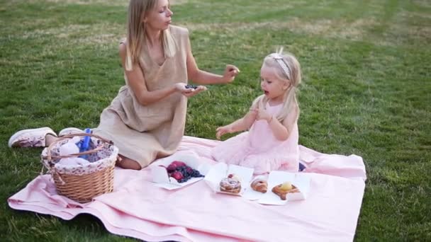 Uma jovem loira e sua filha estão comendo mirtilos. Uma menina e sua mãe estão descansando sentados no gramado em um parque público em uma noite de verão — Vídeo de Stock