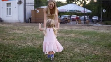 Pembe elbiseli güzel sarışın bir kız çimüzerinde annesinin peşinden koşar. Yetişkin bir anne parkta onunla oynuyor. Annem bebeği kollarına alır ve yere geri koyar. Annem onu yakaladı.