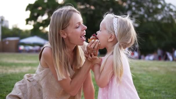 El retrato de la madre y la hija pequeña, pasa el tiempo juntos en el parque de la ciudad en el picnic - muerden el pastel poco de los lados juntos. Mujer joven y niña comiendo dulces, sentados en un — Vídeo de stock