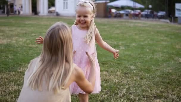 Una hermosa chica rubia con un vestido rosa corre detrás de su madre en el césped. Una madre adulta juega con ella en el parque. Mamá toma al bebé en sus brazos. Madre atrapó a su hija — Vídeo de stock