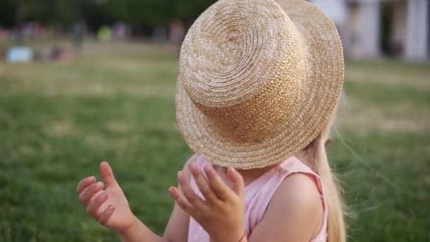Το συναισθηματικό χαρούμενο κορίτσι με το ψάθινο καπέλο να γελάει και να χαμογελάει. Κρύβει το πρόσωπό της πίσω από το καπέλο. Νεαρό θηλυκό παιδί απολαμβάνει το καλοκαίρι και ηλιόλουστη μέρα στο δημόσιο, πράσινο πάρκο — Αρχείο Βίντεο