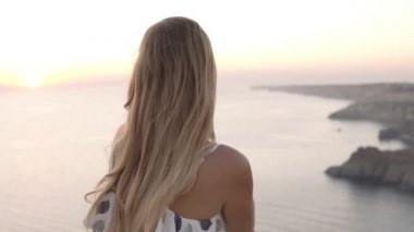 Güzel sundress uzun saçlı genç sarışın kız deniz tarafından kaya üzerinde oturuyor, gün batımı hayran, omzunun üzerinden bakıyor. Arka görünüm