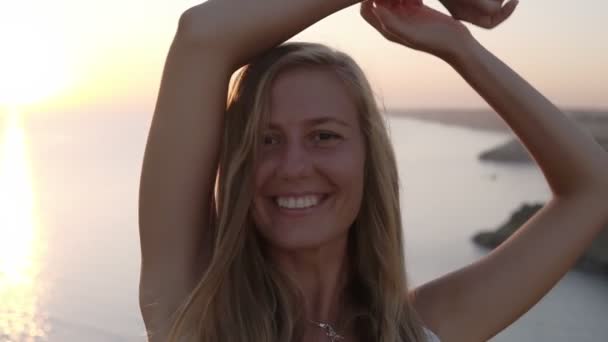 Precioso retrato de una joven rubia parada en el acantilado y sonriendo a la cámara. Chica despreocupada y alegre en la luz del atardecer con las manos extendidas. Hora de verano. Mar en el fondo — Vídeos de Stock