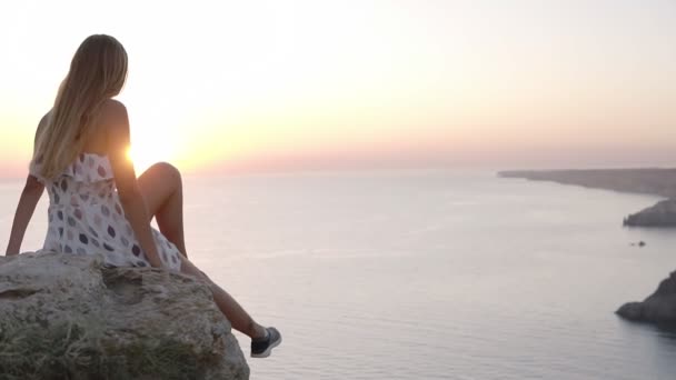 Hermosa joven en vestido bonito está sentada en la roca junto al mar, admirando la puesta de sol, extender las manos en los lados. Vista trasera — Vídeos de Stock