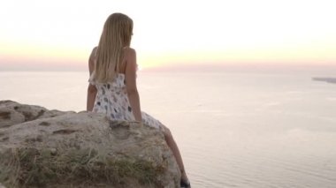 Güzel sundress çekici genç kız deniz tarafından kaya üzerinde oturuyor, gün batımı hayran, kaygısız bacak sallayarak ve omzunun üzerinde bir süre döner. Arka görünüm