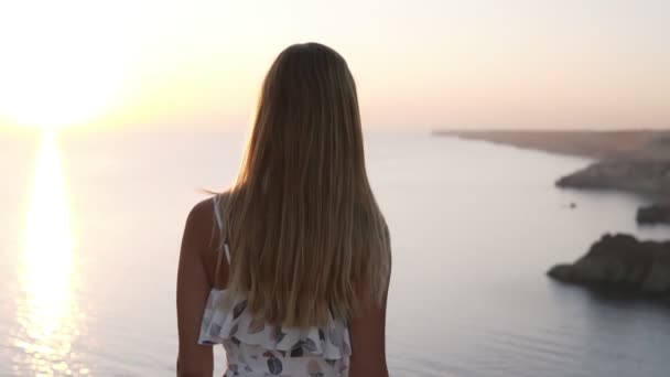 Close back view of fair hair woman tender movement in the air in slow motion. Blonde woman walking by the cliff. Looking on the sea on horizon in sunset and outstretches her hands. Carefree. Slow — Stock Video