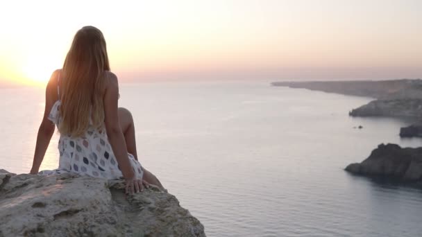Langharig, slank jong meisje in mooie sundress zit op de rots aan de zee, bewondert de zonsondergang, zwaaiende zorgeloos haar been en kijkt omhoog naar de heldere lucht. Achteraanzicht — Stockvideo