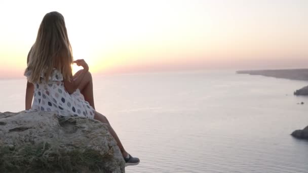 Hermosa joven en vestido bonito está sentada en la roca junto al mar, admirando la puesta de sol, agitando su pierna despreocupada y tocando el largo cabello rubio. Vista trasera — Vídeos de Stock