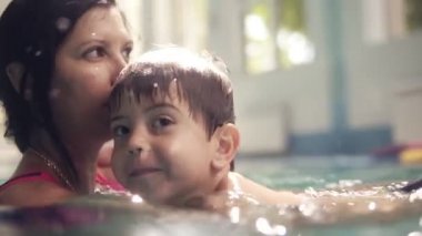 Gülümseyen esmer çocuk annesiyle birlikte havuzda yüzüyor. Onu tutuyor ve yüzmeyi öğretiyor. Aile birlikte vakit geçiriyor. Su altında çekim nasıl çocuk bacakları ile satır