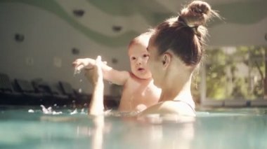 Mutlu genç anne havuzda bebeğiyle oynuyor, suya sıçratıyor. Çocukların gelişimi. Kapalı yüzme havuzunun pencerelerinden parlak ışık yanar. Yavaş çekim
