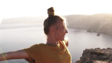 Mutlu, sarı t shirt gülümseyen kadın kamp ellerini yayılır ve sabah güneşi ağırlıyor. Sabah güneş ışığına karşı bir tepede ayakta, uyandım. Arka planda deniz kenarı ve dağlar