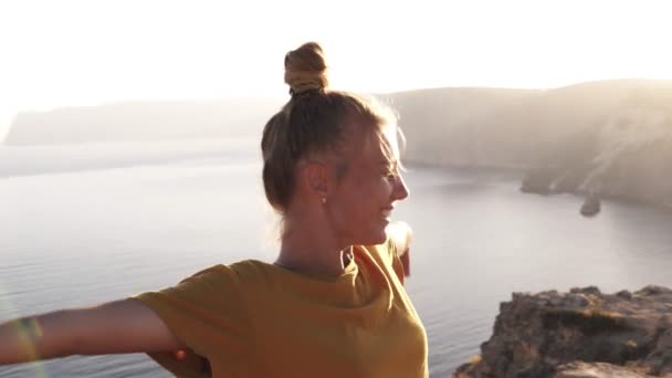 Η χαρούμενη, χαμογελαστή γυναίκα κατασκηνωτής με κίτρινο μπλουζάκι απλώνεται στα χέρια της και καλωσορίζει τον πρωινό ήλιο. Όρθια, στέκεται σε ένα λόφο με το πρωινό ηλιακό φως. Θάλασσα και βουνά στο παρασκήνιο — Αρχείο Βίντεο