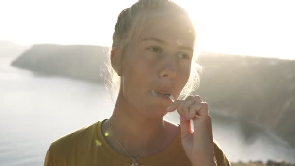 Blonde girl brushing teeth while standing outdoors on the hill with a lake and hills on the background. Traveler, camping concept. Sun shines on the background — Stock Video