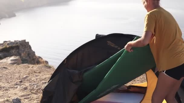 Une femme en T-shirt jaune installant ou retirant un matelas d'une tente de camping. Des images du haut de la falaise. Camping, concept voyageur — Video