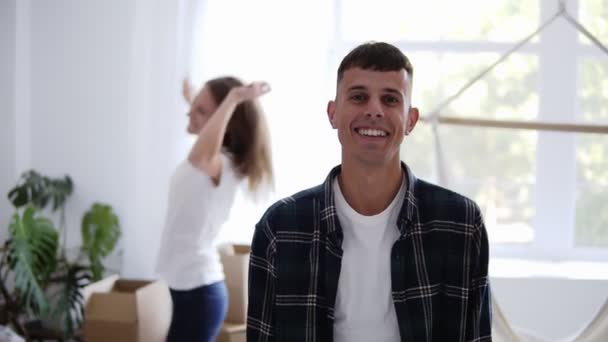 Щасливий білий чоловік посміхається в новій квартирі в сповільненому темпі, поки його дівчина танцює на фоні. Концепція будинку і в новій квартирі — стокове відео