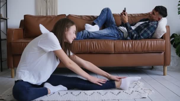 Happy lugn familj fritid hemma Concept, man koppla av på skinnsoffa i komfort vardagsrum med sin smartphone medan hans hustru åtnjuter stretching på varmt golv på mattan. Vy framifrån — Stockvideo