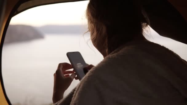 Camping woman is sitting in tent watching taken photos of nature on her smart phone. Camping girl relaxing on vacation. Sea or lake from the top of a hill on background — Stock Video