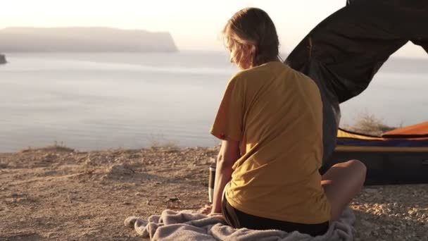 Imágenes de vista trasera de una encantadora chica rubia sentada en el suelo en camiseta amarilla y poniendo una olla con su desayuno campistas al aire libre en el quemador. Camping, estilo de vida salvaje — Vídeo de stock