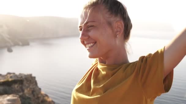 Retrato de campista femenina en camiseta amarilla extiende sus manos y da la bienvenida al sol de la mañana. Despertó, de pie en una colina contra la luz del sol de la mañana. Mar y montañas en el fondo — Vídeos de Stock