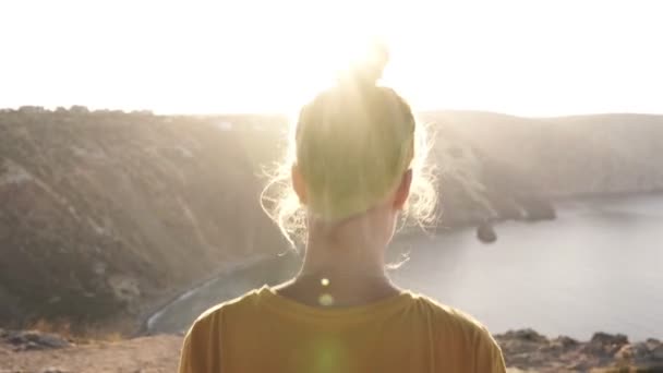 Lento movimento de volta vista lateral do turista feminino caminhando até a borda da montanha e desfrutando de vista sob o penhasco com lago ou mar e colinas, luz solar. Países e conceito de natureza — Vídeo de Stock