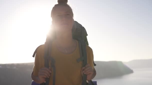 배낭을 가진 젊고 평온한 소녀가 산을 배경으로 산호수 꼭대기를 따라 걷고 있습니다. 여름, 화창한 날, 소녀는 절벽에서 전망을 즐긴다. 해변, 렌즈 플레어. 앞 — 비디오