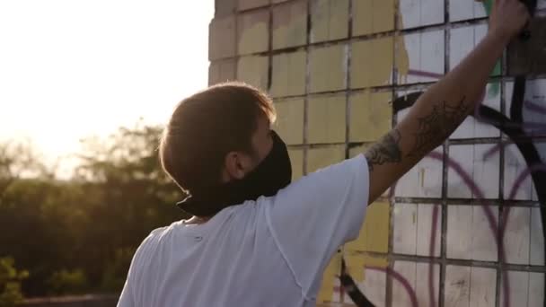 Kaukázusi srác készül a falon a graffiti, festmény graffiti az elhagyott épület görgős festék a bézs színű. Az ember visel alkalmi ruhák és fekete sálat az arcon — Stock videók