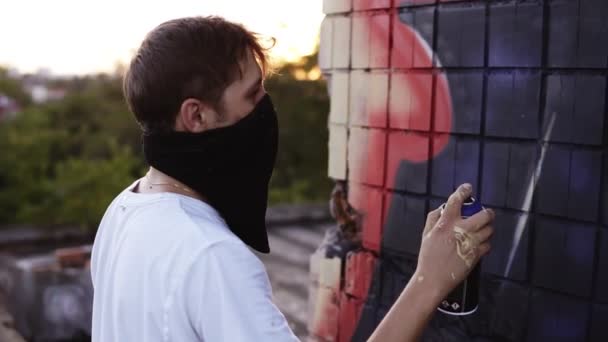 Imágenes de un joven con una camiseta blanca y una bufanda negra en una cara junto a una pared con un graffiti y continuando pintando el cuadro con un spray de color negro. Vista trasera. Al aire libre — Vídeo de stock