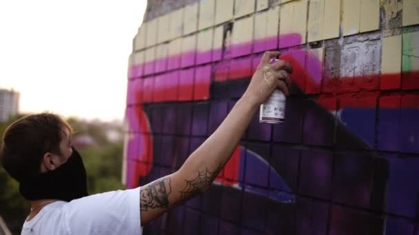 Malířka graffiti s aerosol. Muž v balaklavové a bílé tričko s lahví na spreji. Mladý městský malíř s zakrytou tváří kreslením barevných graffiti na městské ulici v létě na