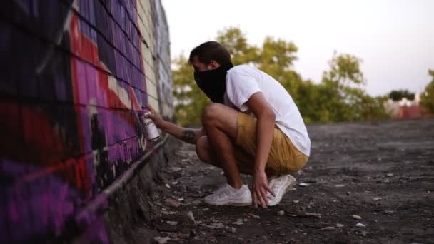 Um tipo sentado em agachamentos a pintar graffiti na parede com um aerossol, vista lateral. Talentosos jovens grafiteiros a pintar. Um homem a desenhar graffiti na parede. Pintor masculino realizando arte de rua no telhado superior — Vídeo de Stock