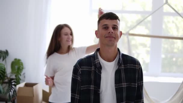 Gelukkig blanke man glimlachend in nieuwe flat in slow motion terwijl zijn vriendin danst op de achtergrond dan springen op zijn schouders, samen lachen. Begrip thuis en in een nieuwe flat — Stockvideo