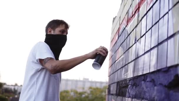 Graffiti művész festés aeroszollal. Férfi símaszkban és fehér pólóban, flakonnal. Lassú mozgás. Fiatal városi festő fedett arccal rajz színes graffiti a városi utca falán a