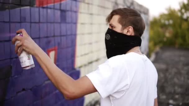 Vista lateral de un joven caucásico en camiseta blanca y bufanda negra en una cara aplicando la pintura azul usando un aerosol en una superficie de edificio al aire libre. Hobby, vandalismo, concepto de arte — Vídeo de stock