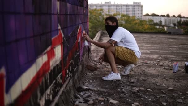 Kerl, der mit einem Spray Graffiti an die Wand malt, Seitenansicht. talentierte junge Graffiti-Künstler malen. Mann zeichnet Graffiti an Wand. Malermeister zeigt Street Art auf dem Dach der Stadt — Stockvideo