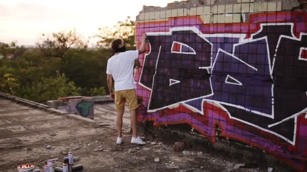 Egy fiatal férfi egy spray-vel be tudja fejezni rajzolni egy új graffiti a falra. Videó arról, hogyan rajzolnak egy graffitit egy téglafalra a tetőn. Lassú mozgás festék alkalmazás a fehér vonalak szóban — Stock videók