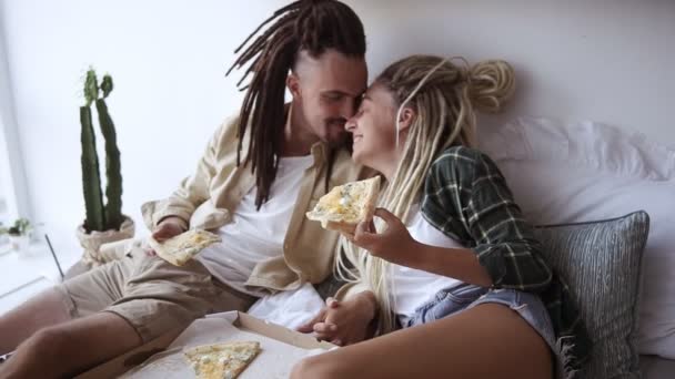 Joven pareja dulce se divierte en el dormitorio, comer pizza de parto, alimentarse, acostarse en la cama, pasar el fin de semana juntos por la mañana. Chicos caucásicos con rastas. Movimiento lento — Vídeos de Stock