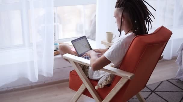 Задние кадры человека в белой футболке с темными дредами, работающего на ноутбуке из дома, сидящего на удобном кресле в гостиной в переднем окне с занавесками. Фрилансер делает свою работу — стоковое видео
