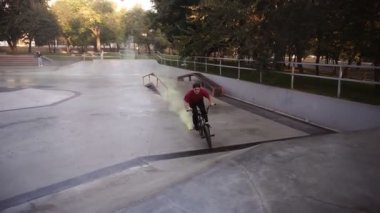 Sarı duman bombası arka tekerleklerde yanarken, boş kaykay parkında pratik yapan beyaz bir BMX sürücüsü. Adam numara yapıyor, bisiklete biniyor ve eğleniyor. Yavaş çekim
