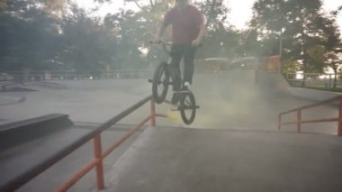 Beyaz bmx binicisi, boş kaykay parkında pratik yapıyor, korkuluklarda kayarken sarı duman bombası arka tekerleklerde yanıyor. Adam numara yapıyor, bisiklete biniyor ve eğleniyor. Yavaş