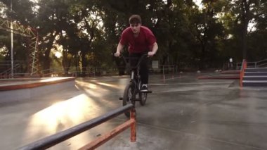 Aktif, genç bir adam siyah bmx motoruyla korkuluklardan atlıyor ve ağır çekimde kayıyor. Kaykay parkında pratik yapan bir adamın yakın çekimi. Gündüz vakti, mercek fişekleri