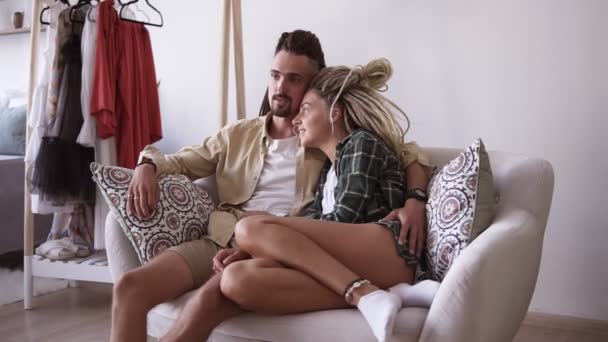 Modern couple sitting embracing on a small white sofa and talking to each other. Stylish couple with brown and blonde dreadlocks, loving people spending quite time together in their own apartments — Stock Video