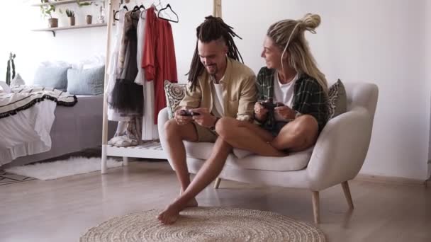 Lustige junge Paar Freund und Freundin spielen Videospiel mit drahtlosen Steuerknüppeln sitzt auf einem weißen Sofa im Wohnzimmer zu Hause. emotionale Menschen genießen Computerspiel, machen — Stockvideo